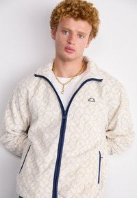 Chaqueta de forro polar crema con textura estampada, cremallera y detalles en azul marino, con un pequeño logo. Incluye cuello levantado y bolsillos laterales.