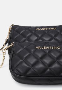 Černé prošívané kožené kabelky se zlatým nápisem „Valentino“. Mají řetízkový popruh a zapínání na zip, s texturovaným povrchem a zakřiveným tvarem.