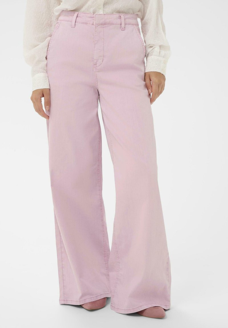Femme portant un pantalon large rose pâle et un chemisier blanc à manches longues, debout les mains détendues le long du corps sur un fond blanc.