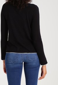 Schwarzes Langarmoberteil mit tailliertem Design, getragen mit blauen Jeans. Das Oberteil hat einen weißen Saum entlang dem unteren Rand.
