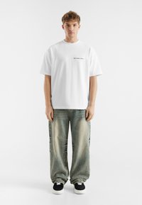 Witte katoenen t-shirt met zwarte tekst "verspild niet beiden", gecombineerd met lichtgewassen, losse jeans en zwarte sneakers. Effen achtergrond.