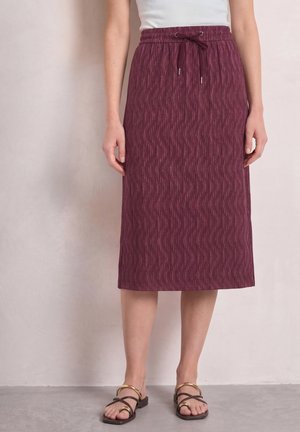Femme portant une jupe midi texturée couleur bordeaux avec une taille à cordon et des sandales noires à brides dorées sur un fond uni.