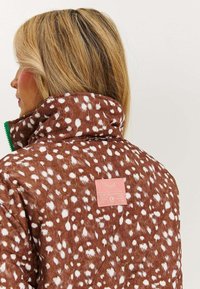 Brązowa pikowana kurtka w białe kropki, z wysokim kołnierzem i zielonym zamkiem. Posiada różową metkę z tekstem na plecach. Miękka tekstura materiału.