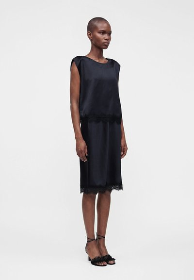 Femme debout portant une robe noire sans manches en satin avec un bord en dentelle à l'ourlet et aux chevilles, chaussée de sandales noires à talons hauts et brides, sur fond blanc.