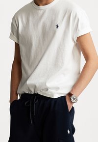 Vit bomull t-shirt med uppvikta ärmar och en liten blå logotyp på bröstet, i kombination med mörka träningsbyxor och en brun klocka.