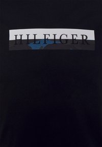 Czarny bawełniany t-shirt z poziomym białym paskiem z napisem "HILFIGER" na tle kamuflażu w odcieniach niebieskiego i szarości.