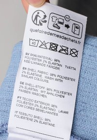 Etichetta di cura dei tessuti con simboli di lavaggio, stampata in più lingue. Composizione del tessuto: 98% poliestere, 2% elastan. Sfondo azzurro chiaro.