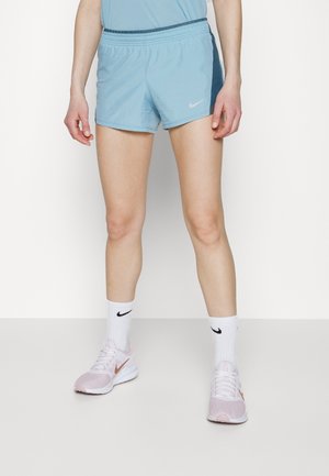 Osoba ubrana w jasnoniebieskie krótkie spodenki sportowe, pasujący top, białe skarpety Nike i jasnoróżowe buty do biegania Nike stojąca na białej podłodze.
