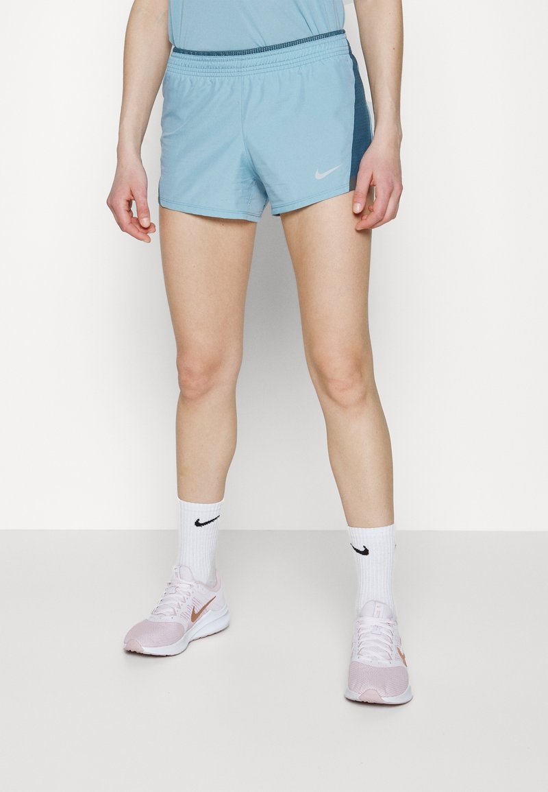 Personne portant un short de sport bleu clair, un haut assorti, des chaussettes blanches Nike et des chaussures de course Nike rose clair, debout sur un sol blanc.