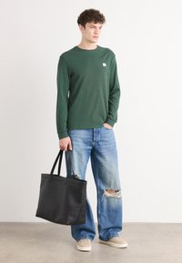 Groene langwerpige shirt met textuur, blauwe denim jeans met versleten details en zwarte tote bag. Casual schoeisel maakt de outfit compleet.