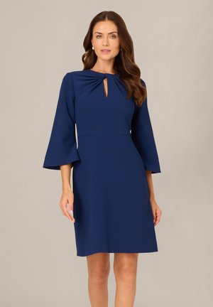 Frau trägt ein knielanges marineblaues Kleid mit 3/4 Glockenärmeln und einem gerafften Schlüsselloch-Ausschnitt, steht vor einem schlichten Hintergrund.