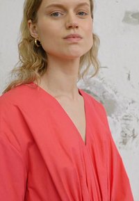 Vestido coral com decote em V, textura fluida, mangas curtas e detalhe franzido. Brincos de argola dourados visíveis, contra um fundo neutro.