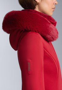 Piros dzseki, melynek kapucnija plüssbéléssel és szőrmével bélelt, anyaga texturált, bal karján RECCO logóval. Testhez álló szabás, strukturált varrásokkal.