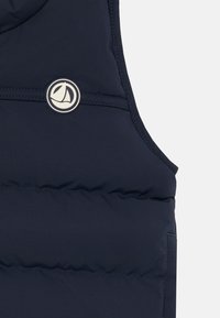 Marinblå puffer-väst med en slät yta, försedd med en rund logopatch. Designen inkluderar en hög krage och en lätt, vadderad struktur.