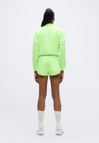 Άτομο στέκεται γυρισμένο με την πλάτη, φοράει μακρυμάνικη μπλούζα νέον πράσινου χρώματος και σορτς, λευκές κάλτσες και λευκά αθλητικά παπούτσια, με απλό φόντο.