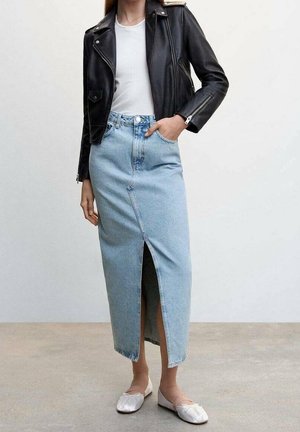 Femme portant une veste en cuir noire, un haut blanc, une jupe en jean bleu clair taille haute avec une fente à l'avant, et des ballerines blanches sur un sol gris.