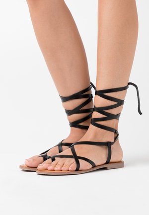 Sandales gladiateur en cuir noir avec une conception à fines lanières, semelle plate et enroulements autour de la cheville, mettant en avant un style minimaliste.