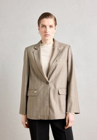 Brauner Nadelstreifenblazer mit taillierter Passform, einreihiger Knopfleiste und zwei Fronttaschen; getragen über einem hellen, gerippten Oberteil.