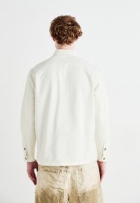 Vit långärmad skjorta med krage, med knäppningar vid ärmsluten och en slät yta, bärs över ljusbeige byxor.