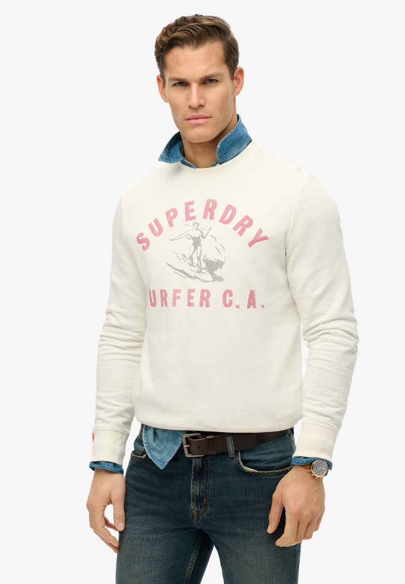 Felpa color crema con grafica "Superdry Surfer C.A.", abbinata a una camicia di jeans con colletto, jeans scuri e una cintura marrone. Texture liscia.