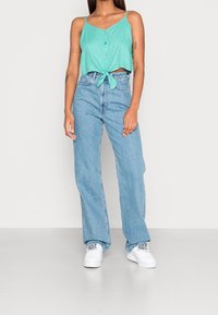 Top corto verde menta con bottoni e nodo davanti, abbinato a jeans a vita alta blu chiaro e sneaker bianche con accenti decorati.