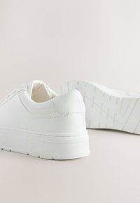 Baltos sportbačiai iš tekstūruotos sintetinės odos su storu padu, su raišteliais ir subtiliais siuvimo elementais.