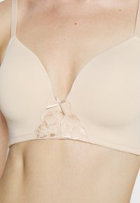 Beige bra with soft fabric, plunging design, lace detailing at the center, and adjustable straps. Smooth texture with delicate accents.