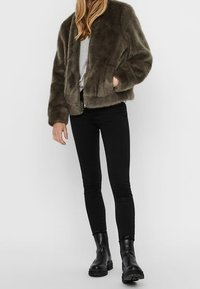 Kvinde iført en mørkebrun imiteret pelsjakke, grå bluse, sorte skinny jeans og sorte ankelstøvler, stående foran en ensfarvet lys baggrund.