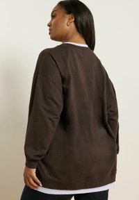 Braunfarbener Langarmpullover mit lockerer Passform und gerippten Bündchen, ausgestattet mit einem geschichteten weißen Saum am unteren Ende und minimalistischen Nähdetails.