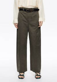 Pantaloni larghi verdi realizzati in misto cotone, con vita alta, passanti per cintura e dettagli cuciti, abbinati a una cintura nera e sandali intrecciati.
