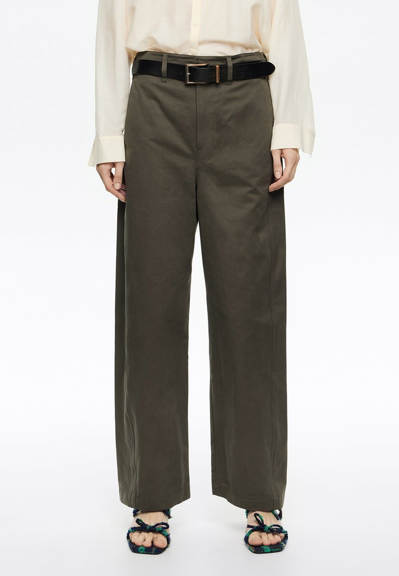 Pantaloni larghi verdi realizzati in misto cotone, con vita alta, passanti per cintura e dettagli cuciti, abbinati a una cintura nera e sandali intrecciati.