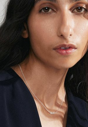 Silverhalsband med "Mom"-hängsmycke som visas på en persons hals, mot en mörk blus. Mjuk textur och delikat kedjedesign.