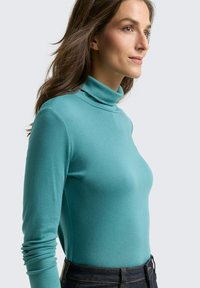 Türkises Rollkragenoberteil aus einem weichen, dehnbaren Stoff, mit langen Ärmeln und einer engen Passform, kombiniert mit dunklen Jeans.