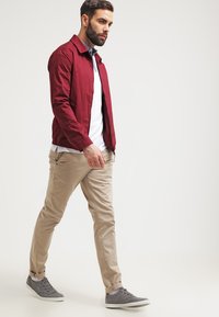 Burgunder lett jakke, hvit skjorte, lysebeige bukser, grå sneakers. Glatt tekstur, glidelås lukking, synlige sidelommer.