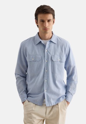 Joven con camisa azul claro de manga larga abotonada sobre una camiseta blanca y pantalones beige, manos en los bolsillos, fondo neutro.