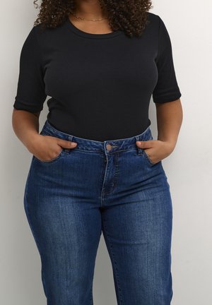 Kvinde med krøllet hår iført en sort top med korte ærmer og højtaljede blå jeans med hænderne i lommerne foran en ensfarvet baggrund.