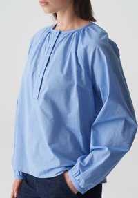 Blusa azzurra chiara a maniche lunghe con scollatura tondeggiante, dettagli plissettati attorno alle spalle e polsini elasticizzati, realizzata in un tessuto liscio.