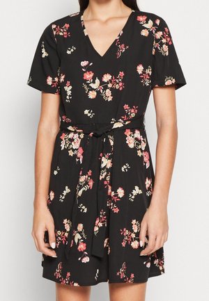 Femme portant une robe noire à manches courtes avec un imprimé floral rose et blanc et une ceinture à nouer à la taille.
