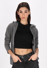Felpa grigia con zip realizzata in un tessuto morbido, indossata sopra a un crop top nero aderente. Combinata con jeans neri a vita alta e una cintura nera.
