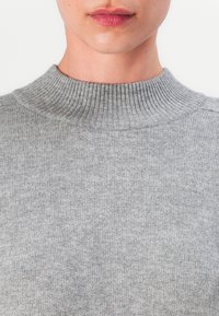 Hellgrauer Strickpullover mit hohem ribbeligem Halsausschnitt und weicher Textur, der ein einheitliches Muster über den gesamten Stoff aufweist.