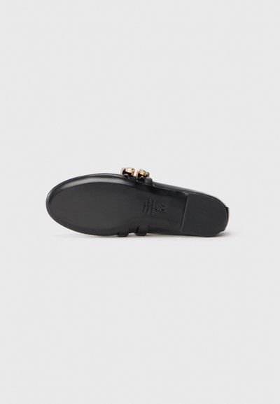Svart sandal med platt sula, med en slät yta och två dekorativa spännen i guldton. Minimalistisk design som betonar elegans.