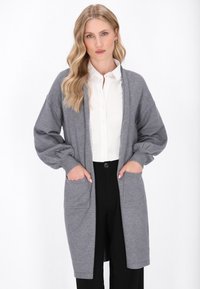 Grauer Strickcardigan mit langen Ärmeln, lockerer Passform, seitlichen Taschen und gerippten Bündchen, kombiniert mit einem weißen Hemd mit Kragen und schwarzen Hosen.