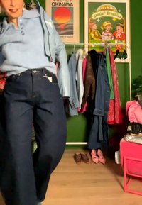 Person mit hellblauem Hoodie und dunkler Hose steht in der Nähe eines Kleiderständers mit Jacken und Kleidern, mit sichtbaren Postern und Schuhen.