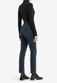 Jersey de tortuga negro acanalado combinado con jeans de cintura alta de color azul oscuro. Las botas tienen un tacón grueso. El fondo blanco destaca el conjunto.