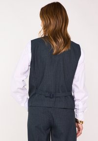 Gilet a righe sottili scuro realizzato in tessuto strutturato, caratterizzato da una cintura sul retro, indossato sopra una camicia bianca con bottoni sui polsini.
