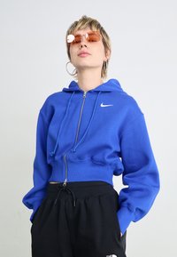 Izgriezta zila kapuci jaka ar priekšu zipu un auklu kapuci. Uz krūšu ir balts Nike logo, apvienota ar melniem sporta biksēm.