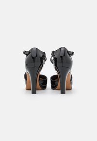 Des chaussures à talons aiguilles en cuir verni noir, avec un bout pointu, une bride autour de la cheville et un talon stilettos de 10 cm, présentant une finition lisse et brillante.