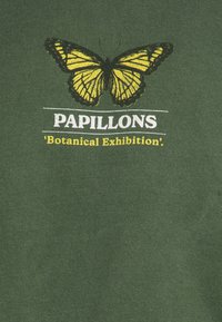 Zelené bavlněné tričko s grafikou žluté a černé motýlice. Text dole zní "PAPILLONS 'Botanická výstava'."