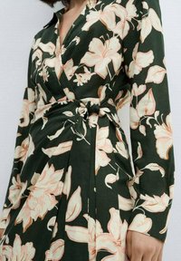 Robe portefeuille à manches longues, avec un fond vert foncé et de grandes lys roses pâles. Se noue à la taille avec un détail froncé.