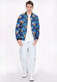 Marine bomberjack met koraalrifpatroon in blauw en oranje, gecombineerd met lichtblauwe cargobroeken en witte sneakers.